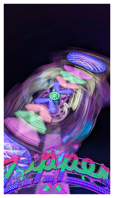 night. timed exposure with motion blur. low angle view of a ride at the county fair. in the foreground, at bottom, the gateway is the word ZIPPER in pink green and purple lights. behind it, a blur of lights in many similar colors against a black night sky.