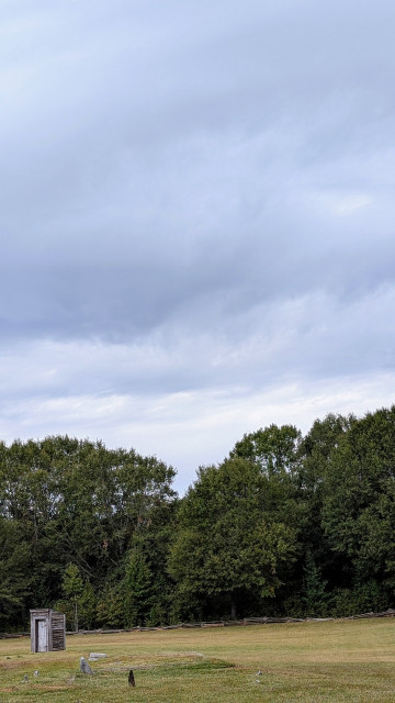 an old wooden outhouse stands in a field with a tree line behind a split rail fence in the distance. A couple of fieldstone grave markers appear in the foreground. The sky above is mostly cloudy.