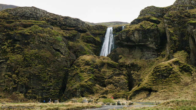 A photo of a rockface under a cloudy sky. The grey rocks are covered in mosses and lichens, and are deeply scored by water and weather erosion. Behind a large boulder, the top of a waterfall is visible. Deep clefts on either side of the huge stone show that people have walked through many times, and a few visitors can be seen standing at the outside of the paths.