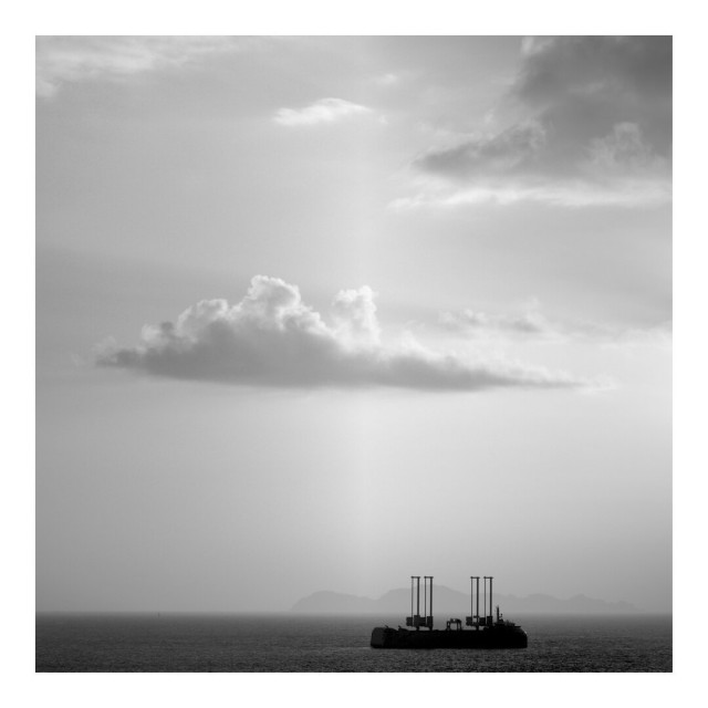 Black and white image of a strange boat in the bottom of the frame, under light and clouds. Low horizon line.