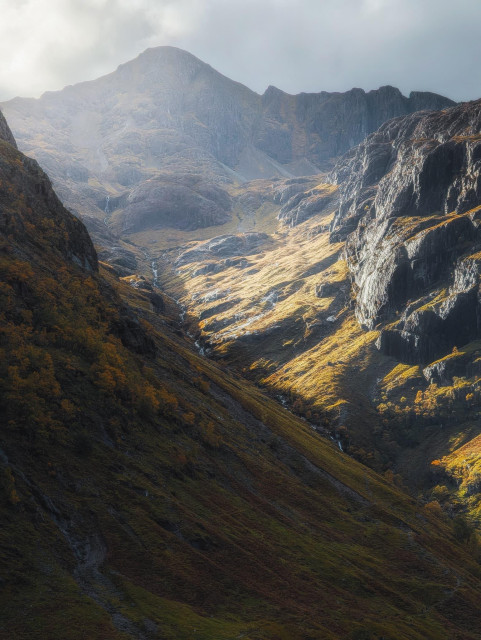 A breathtaking view of Corrie nan Lochan nestled between two of the Three Sisters of Glencoe, showcasing the raw, rugged beauty of the Scottish Highlands. The image captures a dramatic mountain landscape bathed in golden autumnal light. Steep, craggy slopes rise on either side of a narrow glen, their rocky faces etched with shadows. A winding stream cascades down the centre, glinting in the sunlight. The vegetation on the hillsides ranges from rich golden hues to deep browns, indicating the changing seasons. In the background, a majestic mountain peak looms, partially obscured by mist, adding to the ethereal atmosphere. The interplay of light and shadow accentuates the contours of the landscape, creating a sense of depth and grandeur that epitomises the wild, untamed nature of Glencoe.
