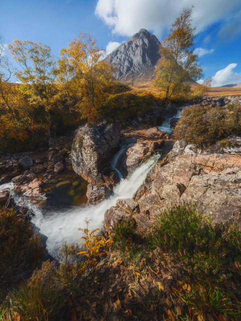 A captivating autumnal scene at River Coupall near Glencoe, with the iconic Buachaille Etive Mor mountain dominating the background. In the foreground, a cascading waterfall tumbles over rugged rocks, its white water contrasting sharply with the surrounding autumn-hued vegetation. Golden-leaved trees frame the scene, their branches reaching towards a vibrant blue sky dotted with wispy clouds. The river winds through the rocky terrain, leading the eye towards the imposing pyramid-shaped peak of Buachaille Etive Mor, its grey, craggy face a stark contrast to the warm colours of the Highland landscape. The interplay of light and shadow across the rocks and foliage creates a rich tapestry of textures, capturing the raw beauty of the Scottish wilderness in its autumnal glory.
