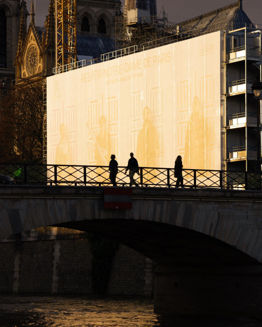 A striking view of Notre Dame Cathedral in Paris under renovation. In the foreground, three silhouetted figures walk across a bridge over the Seine River. The bridge's ornate railing is visible. Behind them, a large illuminated construction screen covers part of the cathedral, reading 'REBATIR NOTRE DAME DE PARIS' (Rebuilding Notre Dame of Paris). The screen shows a faint outline of the cathedral's architecture. In the background, the cathedral's gothic spires and scaffolding are visible against a dusky sky. The scene is dramatically lit, with the golden light on the screen contrasting with the darker surroundings.