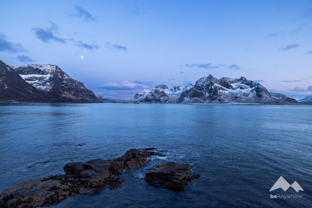 Mountains in the Atlantic Ocean lightly covered in snow. The ocean looks blue, the sky light blue. Moon is rising.