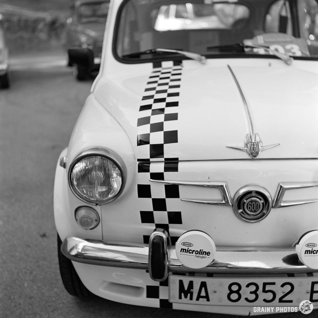 A black-and-white film photo of a classic Fiat 600. The photograph is of the front of the car, a three-quarter view. There is a black and white chequered pattern running along the front grill and bonnet.