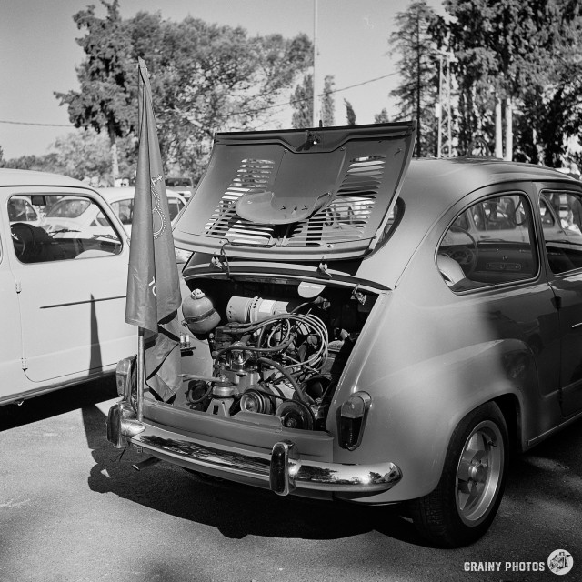 A black-and-white film photo of a Seat 600. The photograph is of the rear of the car with the bonnet open, exposing the engine bay. A Spanish flag is fixed to the rear bumper of the car.