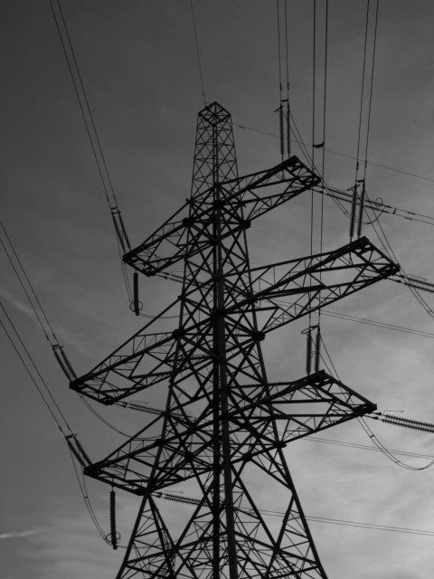 Monochrome photo taken looking up at a large metal frame electricity pylon silhouetted against a gloomy sky. Power cables come in from the direction of the top of the frame and leave at the right.