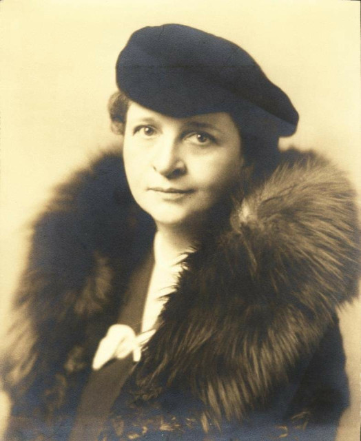 A sepia photo of a demure woman wearing a black beret and a fur collar. She is looking at the camera.  She is wearing a dark dress and a white blouse.