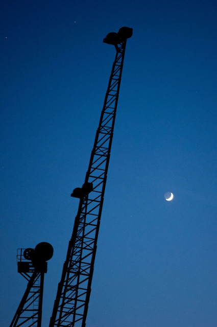 Photo of the top of an upgright dock crane, black latticework and duble pulleys silhouetted against a blue evening sky. At the top left, a handful of stars, at the middle right, a crescent moon