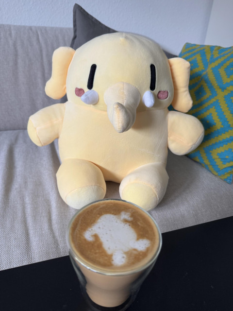 Mastodon Plushie sitting on the couch with a coffee in front. The coffee has latte art that tries to resemble the plushie.