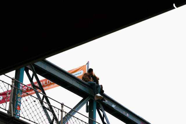 A couple sits intimately on the steel beam of a bridge or elevated structure, their legs dangling over the edge. The structure appears to be teal-colored. Above them, a partial view of an orange banner is visible, with '.freiburg.de' partially legible. The scene is framed by a dark overhang at the top of the image, creating a sense of voyeuristic perspective. Below the couple, parts of the bridge's railing and some colorful signage can be seen.