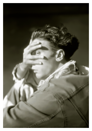 side view of a man with wavy, dark hair in a faded jean jacket and jeans. he's sitting with his left arm in the foreground, almost covering his face with his right hand with a space between the fingers revealing his left eye making contact.