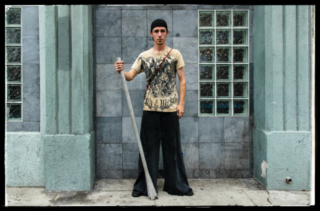 A portait of a young man and his didgeridoo, standing on a sidewalk in front of a jade green building. He is wearing a yellow t-shirt and holding his long wind instrument at his side. Oddly out of context for an American city but this is San Francisco.