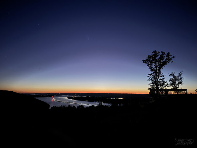 Wide angle view of a big river from a tall overlook bluff. It's so dark everything but the sky and the reflective water is black. Some of the brightest stars are peering out, and in the middle of the sky just peeks out the comet, starting to show as the glow on the horizon turns deep orange.