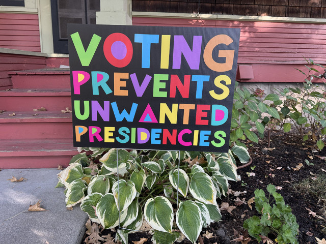 A political sign above some hosta plants says "Voting Prevents Unwanted Presidencies." The letters are a variety of bright colors on a black background.