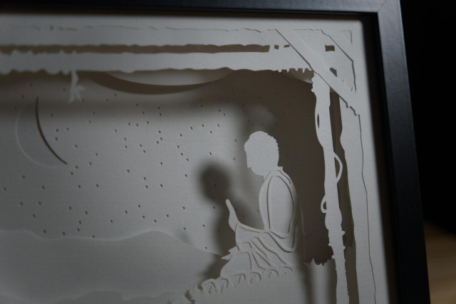 Intricate cut layers of paper stacked in a deep frame. The scene depicted here is of the Tian Tan Buddha in Hong Kong, with a star and moon filled sky.