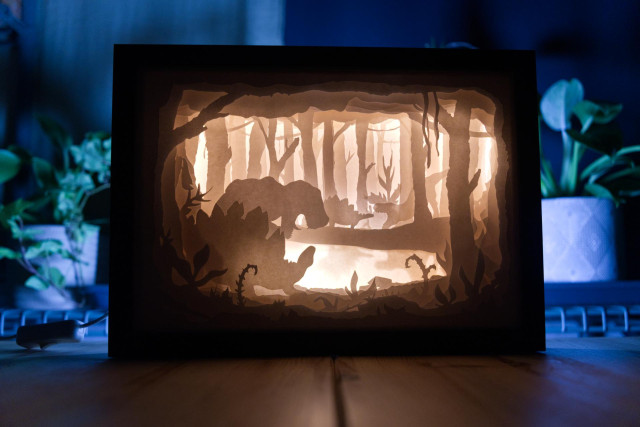 Intricate paper cut design made from multiple layers and placed in a deep frame. The scene depicts dinosaurs in the jungle. It's being backlit by LED.