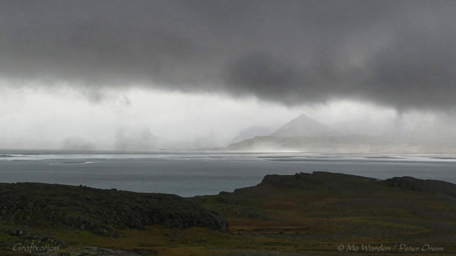 A photo of a craggy coastline in the foreground with a seascape beyond. On the other side of a wide bay is a sharply pointed mountain at the end of a promontory. Thick cloud is hanging overhead, and fog has already crept across the headland in front of the viewpoint. Mist is hanging across the water and continues towards the ocean on the left. The shot is mostly in shades of grey, and is very dramatic.