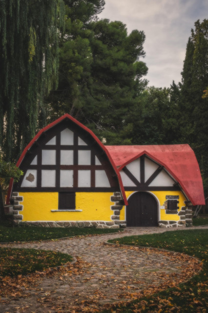 Fotografía vertical donde se ve en la parte central una casita de cuento similar a la de Blancanieves. En la parte inferior un caminito de hojas que lleva hacia ella. En la superior árboles y algo de cielo.