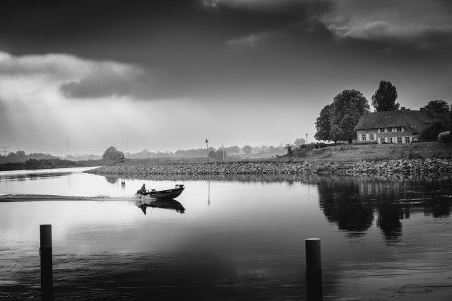 Rivier met een klein bootje dat voorbij vaart. Aan de overkant en rechts van de foto staat een boerderij.