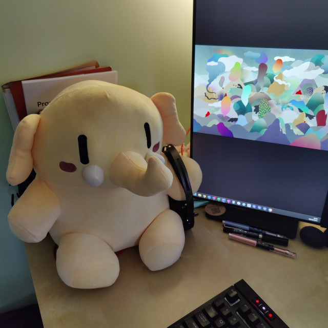 Yellow mastodon plush toy, sitting on a desk, holding a headset, next to a screen with abstract background 