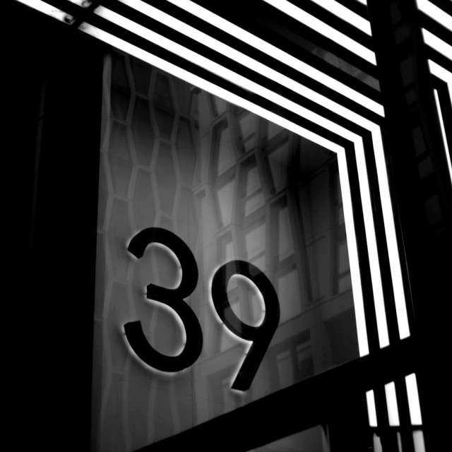 Black and white photo in which a building with windows in an elongated honeycomb pattern is reflected in another window, behind which, on a wall, is a back-lit '39' and which has striking white rectilinear lines angling above and to the right.