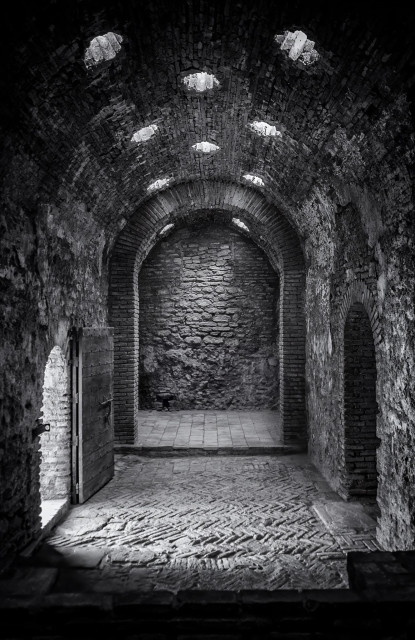 Darkened entryway to Roman bath in Spain with light spilling in from an open door. Built of stone and concrete, the edifice features tall, arched ceilings with vent holes,