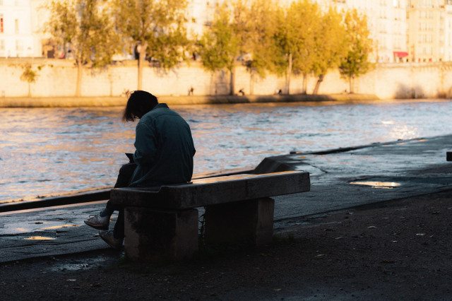 A person sits alone on a concrete bench beside a river, with their back facing the camera. They are hunched over, possibly looking at a phone or a book. The warm light of the setting sun casts a soft glow on the trees and buildings in the background, which are reflected in the gently rippling water. The overall mood feels peaceful and slightly introspective, with the quiet surroundings and late afternoon shadows creating a sense of solitude.