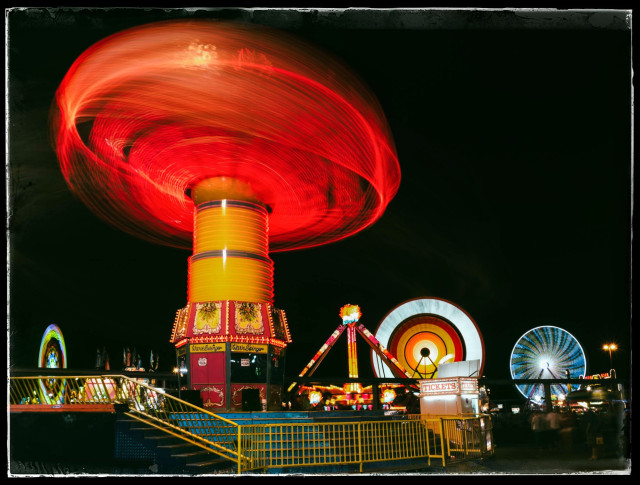 A 6 second exposure of five carnival rides at night. 

Three of the rides, in the distance, are colorful, spinning ferris wheels. With their green, blue, orange, red and white lights, they slowly grind people to bits like grist in a mill.

The main attraction, in the foreground, is a giant, spinning centrifuge. It is blood orange. You can imagine what this does to the riders who  are foolish enough to pay for a ride.