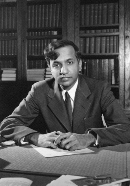 Black and white photo of a young Subrahmanyan Chandrasekhar, seated at a table with shelves full of books behind him. His hands are folded on the table in front of him and he is looking directly at the camera. Chandrasekhar wears a dark suit and tie, with a white, collared shirt.
