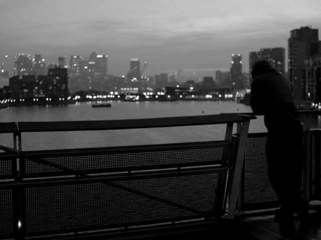 Monochrome photo of a high bridge over a body of water with apartment blocks and then skyscrapers in the background. It's not quite night yet, but clearly late, as there's not lots of contrast in the image, but also because lights are on in all the buildings. In the foreground, on the right, leaning on the bridge railing, is a figure, just about silhouetted against the bridge and a dark block.