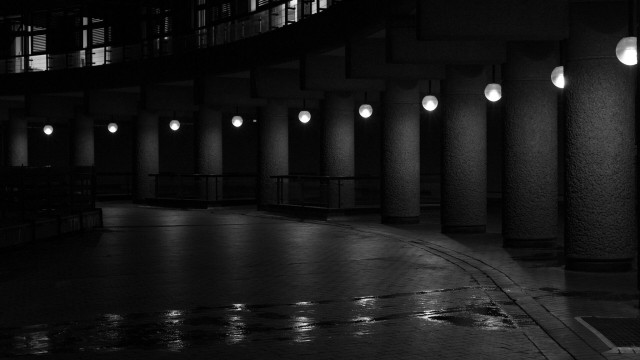 Dark black and white photo of a curve of rough concrete pillars, between each of which is a globe light mounted in the ceiling. In the open space in front, a splash of water reflects their light.