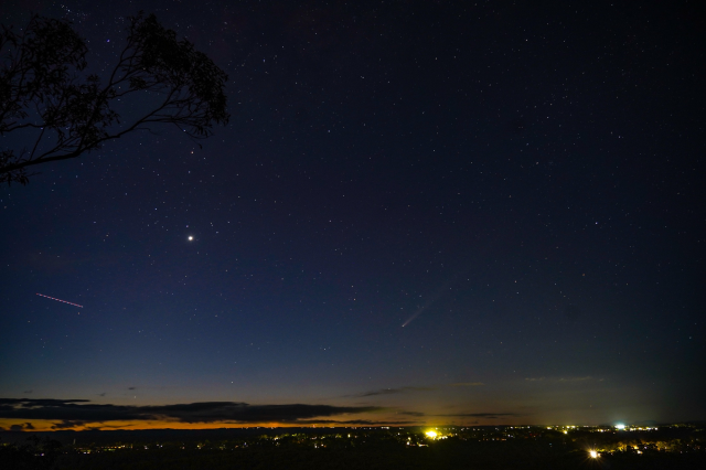 Looking out across the horizon over the city lights, where the night sky is filled with stars, the planet Venus, a faint comet and an aircraft trail. 