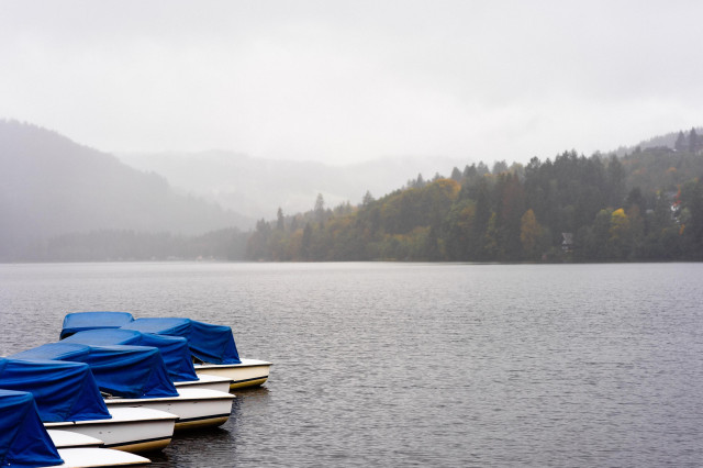 A serene lake scene on a misty day. In the foreground, several small boats with bright blue covers are moored at the shore, their reflections visible in the still water. The lake stretches out to the horizon, where forested hills rise up, their slopes covered in evergreen and deciduous trees showing hints of autumn colors. The entire scene is enveloped in a soft haze, creating a tranquil, dreamlike atmosphere. The muted colors and calm water evoke a sense of peaceful solitude in nature.