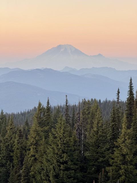 A view of Mount Rainier at sunset on a hazy evening from my campsite on Mount Adams. An orange sky radiates over the prominent volcano. White glaciers on the peak. Tall green trees in the foreground with hazy mountains beyond. Photo taken October 12, 2024, in Washington State, U.S.