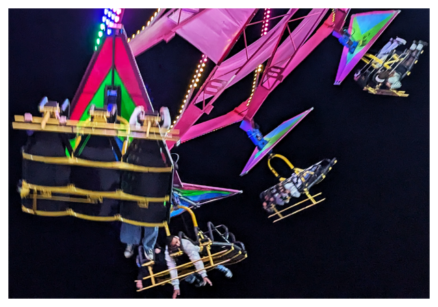 night. 4 rider compartments on a colorful carnival ride as it circles at a 45º  angle above the unseen ground with interesting rider postures in the two foreground compartments. black sky in the background.