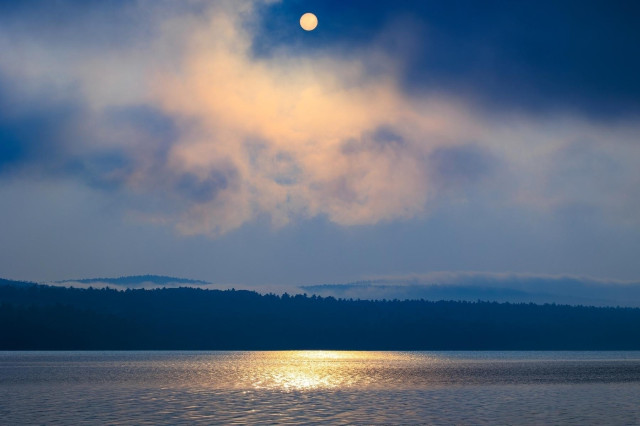 A serene river reflects the soft glow of the setting sun through misty clouds and over distant hills.