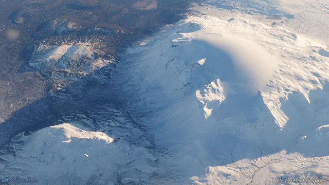 A photo taken through the window of a plane. The ground surface is dark rock which is fairly clear of snow and ice but, in the shelter of the mountains, there are large ice-fields. One flat-topped mesa has a huge dome of snow near the centre, and other smaller mountains are standing by its shoulder. In the lower right, a meltwater river is cutting through the ice in a dark, twisting thread.