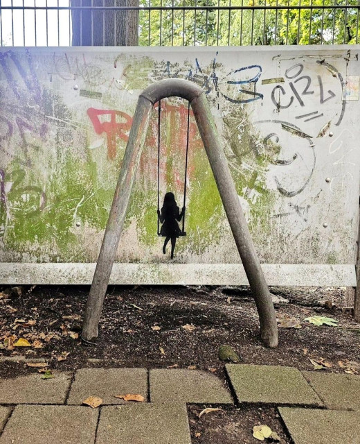 Streetartwall.  A small mural with a girl on a swing was sprayed on an old, dirty street wall. The small mural is sprayed as a black silhouette. A bent metal pipe (like an upside-down "U") stands in front of the wall as a limiter. The little girl hangs between the two pipe bends on long painted ropes and swings. A simple but sweet little mural by the wayside.
