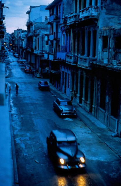 Photography. A color photo of a street view at "blue hour" in Old Havana. In the foreground, a wide, dark American vintage car drives quickly through the darkness with dim lighting. Some vintage cars are parked in front of the houses. The asphalt of the road is crumbling, rivulets of rain run down the sides. On the right are the old multi-storey houses, whose former beauty can still be glimpsed. Occasionally, the silhouettes of people can be made out as they make their way through the city. Nothing lights the way, there is no light from street lamps. Through the cool light and the blue of the night, the photo creates a place that begins to come alive in the imagination.
Info: 
David Alan Harvey (born 1944) is an American based Magnum Photographer. He is one of the living legends in street photography. His photography combines a direct documentary style and emotional mood with his own powerful, personal vision. 