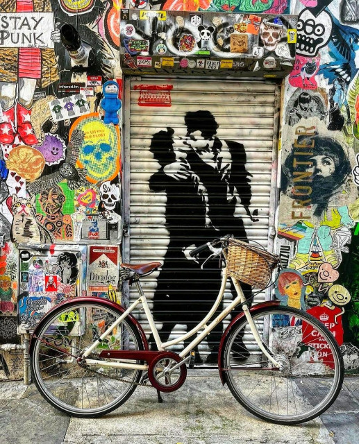 Streetartwall. Next to a street wall with countless stickers and paste-ups for a festival, a large paste-up of a kissing couple has been spray-painted. The black silhouette of a young man in a black suit intimately kissing a woman in a skirt, coat and high heels is sprayed onto a white roller shutter. The photo shows a bicycle in front of the mural, which belongs to the photographer and was placed there especially for the occasion).