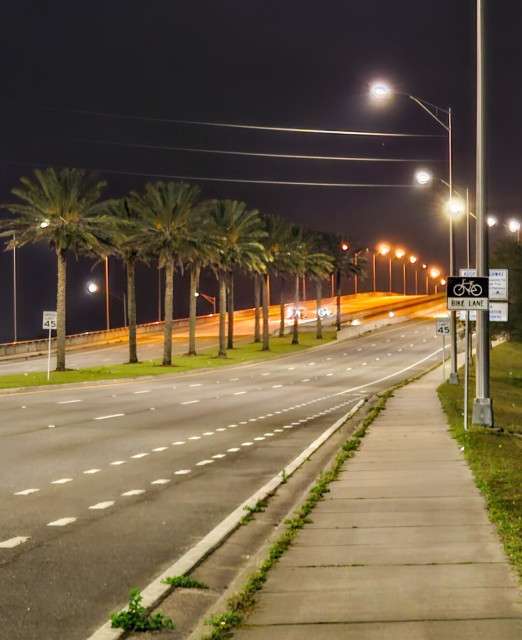 Late night approaching one of my favorite bridges to cross at night. Multiple lanes of illuminated roadway lead to and from the arc of a large concrete bridge ahead. With a green grassy median, lined with palm trees. And pedestrian sidewalks will illuminated with countless street lights, extending far into the night.