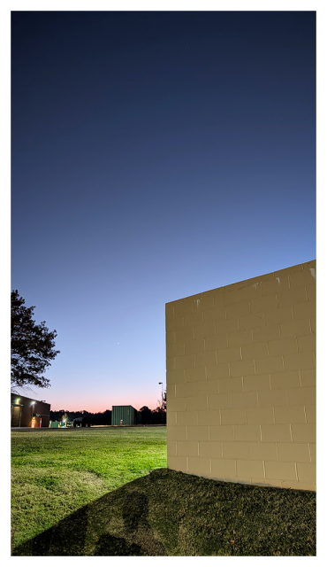 the horizon is pink behind a line of trees in the distance. the sky is shades of gray/blue. in the foreground a beige cement block dumpster enclosure casts a shadow onto green grass where you may also spot my shadow. lit by streetligh,t in front of the structure, in the mid-distance, the right half of a tree appears to hover over single story brick building at left with several lights.