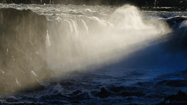 A photo of a waterfall, strongly sunlit from the right. The spray from the falls is rising higher into the air than the crest of the cascade itself. In the background, a powerful river can be seen approaching the drop. The dark rocks on either side are shining with moisture. At the base of the falls, a turbulent river is surging towards the viewpoint and some rapids can be seen. From the top right to the bottom left of the waterfall, strong sunbeams are slicing through the spray.