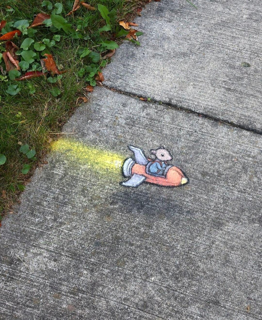 Streetart. A little mouse in a blue dress (called Nadine) and a rocket have been drawn on a sidewalk with chalk. Nadine sits in the red rocket and whirls horizontally across the ground with a long tail of fire. Title: "Nadine Runs a Quick Errand."