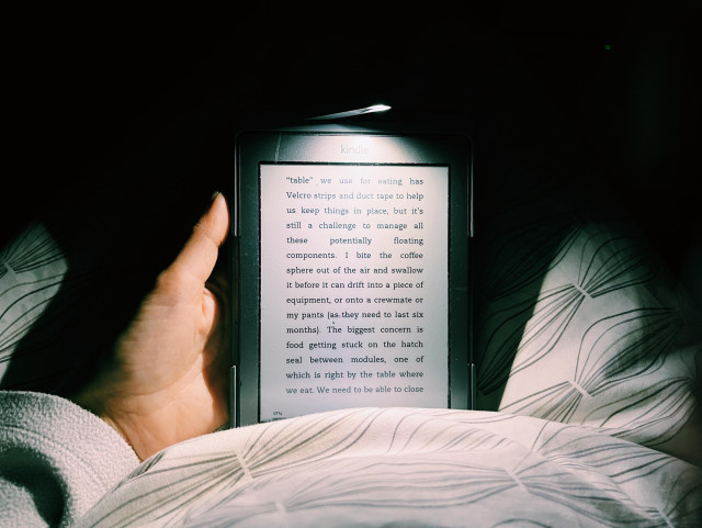 A photograph of a hand holding up a Kindle in the dark, with a built-in light that shows a page from Scott Kelly’s book Endurance