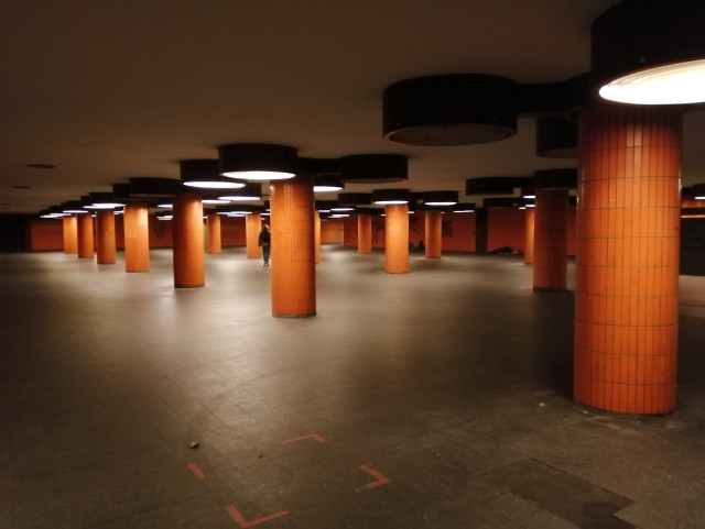 A poorly lit empty subterranean pedestrian area.

It gives you the impression of desolation.

The ceiling is held by round orange columns. Light is provided by round lumenescent circles on the ceiling.

A few homeless people are sleeping near the orange walls. 