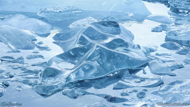 A photo of a water surface softly sunlit from the right, clean turquoise water with odd pieces of ice floating in it. Most of the shards are smoothly sculpted by being rolled in the water as they melted. Some are broken pieces, but even these have rounded edges. Some clear water is visible and a larger, central chunk has a small pool within its curves. Rings on the surface of the water show that it's raining gently. The ice and the water are all a gorgeous shade of cyan.