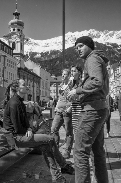 Das Bild ist im Hochformat und in schwarz-weiß fotografiert worden.
4 junge Menschen machen Pause beim Stadtbummel in Innsbruck. Eine junge Frau sitzt auf einer Bank, 3 andere stehen um sie herum (2 Frauen und ein Mann). Man sieht die Bergkulisse der Nordkette, die schneebedeckt ist und den Kirchturm der Spitalkirche.