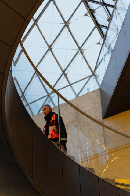 A person wearing a high-visibility orange safety vest and black jacket stands on a curved staircase in what appears to be a modern building. Above them, a dramatic circular skylight with geometric diamond-patterned glazing lets in natural light. The architecture features clean lines with dark walls and yellow accents visible in the lower portion of the frame. The composition creates a striking contrast between the figure and the architectural elements.